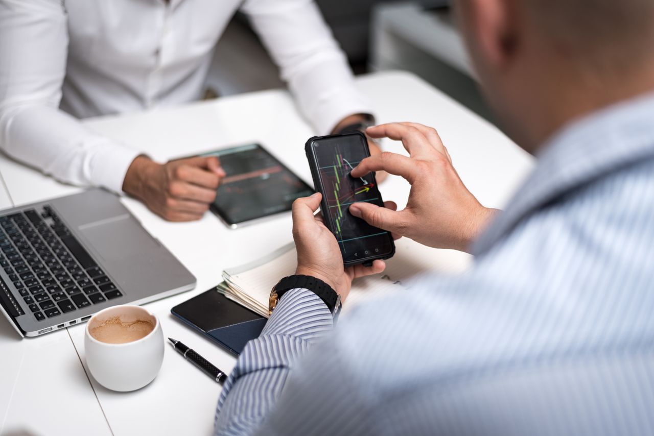 Business professionals using smartphones and tablets to review trading charts during a meeting.